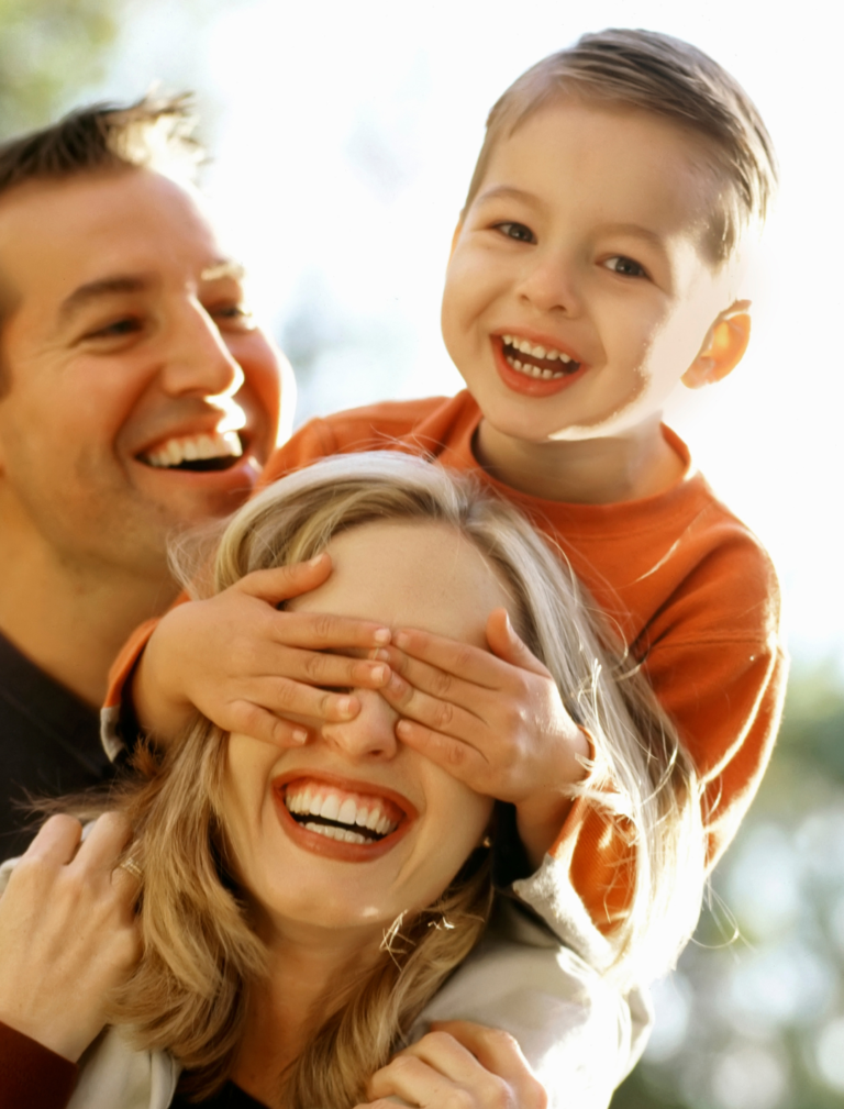 Happy family. Small son is covering smiling moms eyes while sitting her shoulders. Dad is laughing up at son.